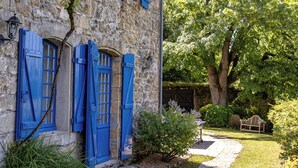 Un edificio de piedra con contraventanas azules, un camino de piedra y un gran árbol al fondo.