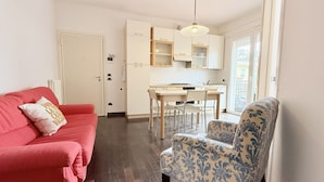 A compact living space with a kitchenette, a red sofa, a patterned armchair, and a door leading to an outdoor area. - RN002 – Rimini, Comfortable Two-Bedroom Apartment Near the Sea with Terrace (Rimini)