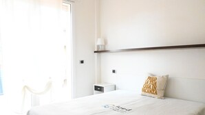 A hotel room with a bed, a bedside table, a nightstand, a lamp, and a window with curtains. - RN002 – Rimini, Comfortable Two-Bedroom Apartment Near the Sea with Terrace (Rimini)