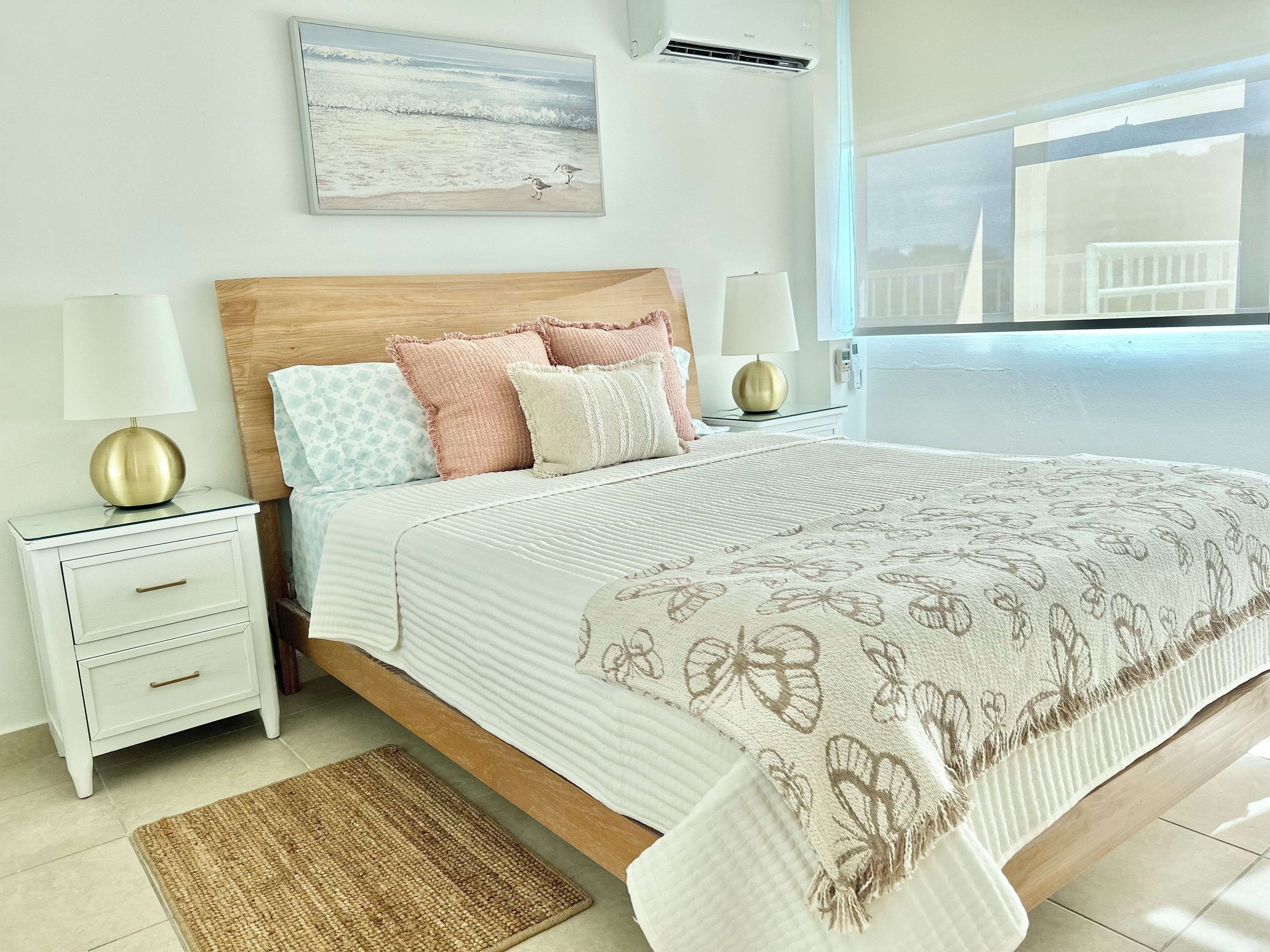 Quarto com cabeceira de madeira, colcha branca com padrão floral, duas mesas de cabeceira com abajures e vista para o exterior pela janela.