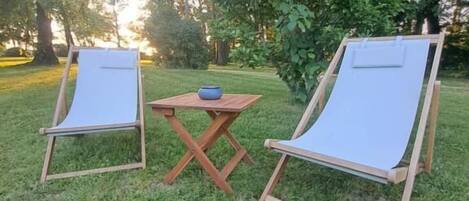 Two wooden lounge chairs with canvas seats and backs, positioned on a grassy lawn with a small wooden table and a blue bowl between them.