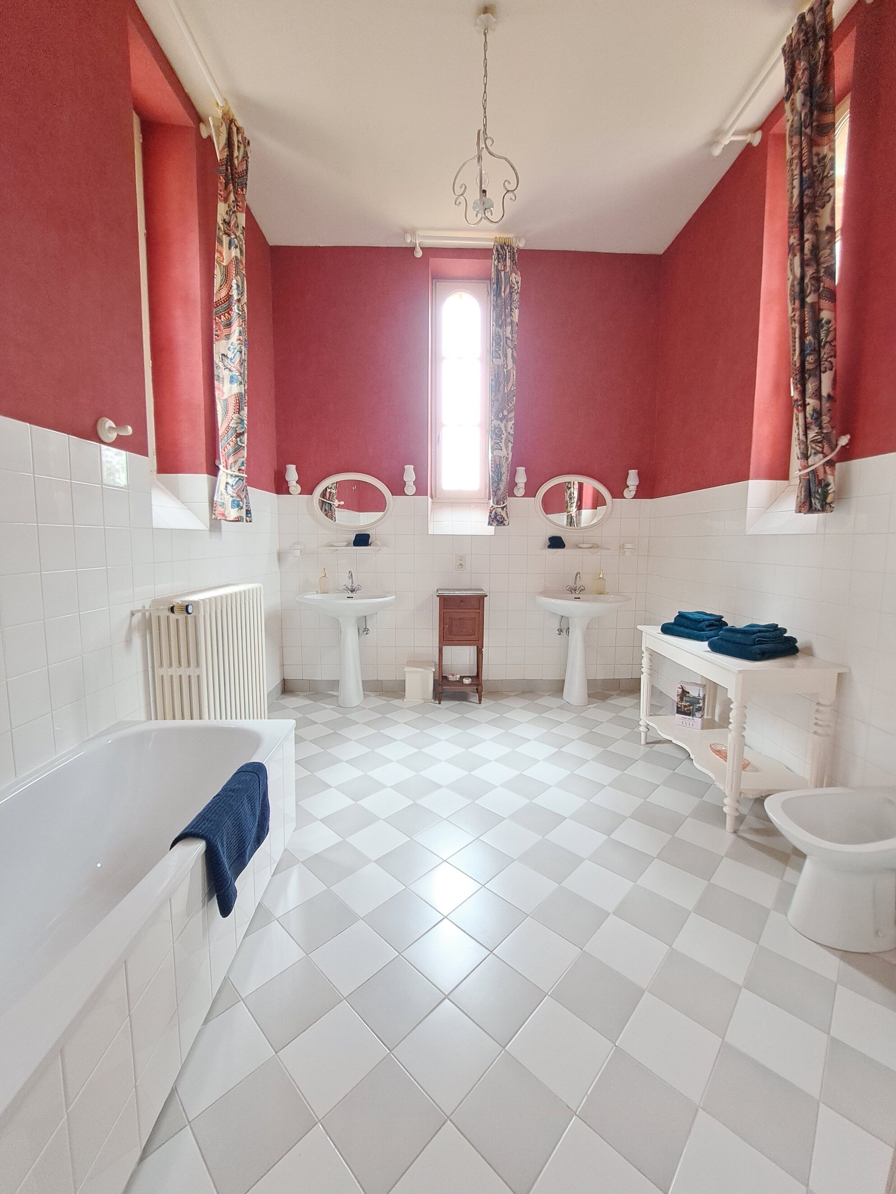 A bathroom with a white bathtub, two sinks, a toilet, a radiator, and a window with curtains.