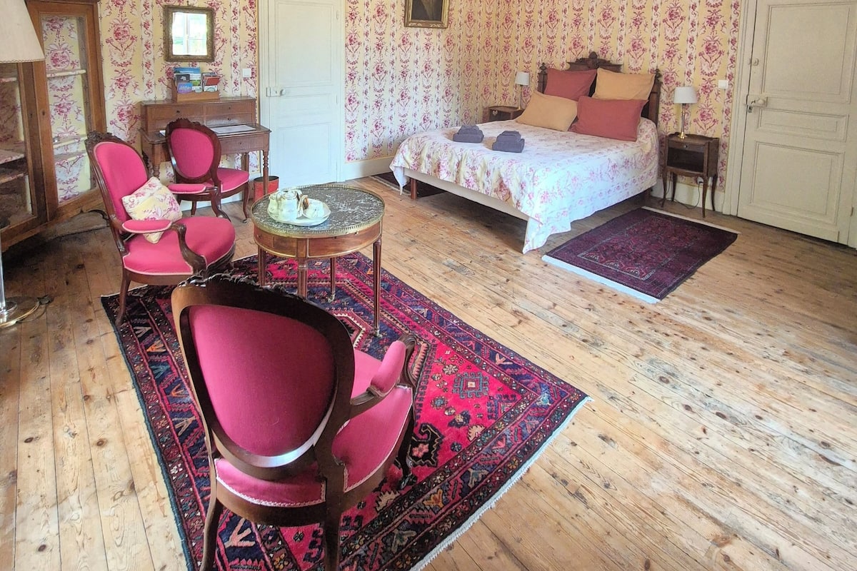 A room with a bed, two chairs, a small table, a wooden floor, and floral wallpaper.