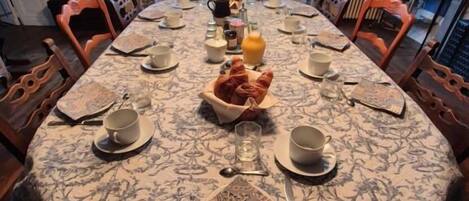 A dining table set for breakfast with a patterned tablecloth, chairs, and a view of the outdoors through large windows.