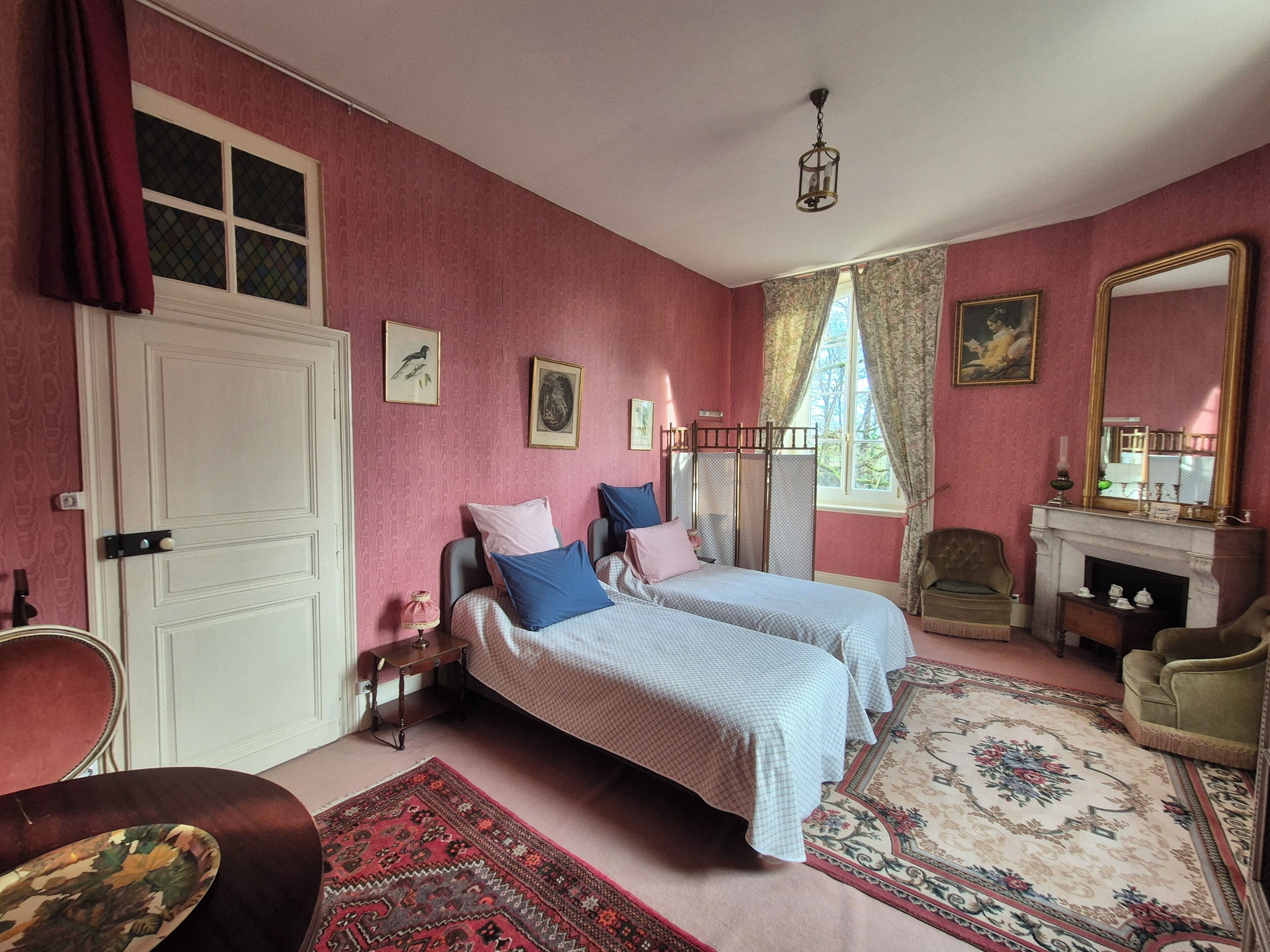 Ein Zimmer mit zwei Betten, einem Sessel, einem Kamin und einem großen Spiegel.