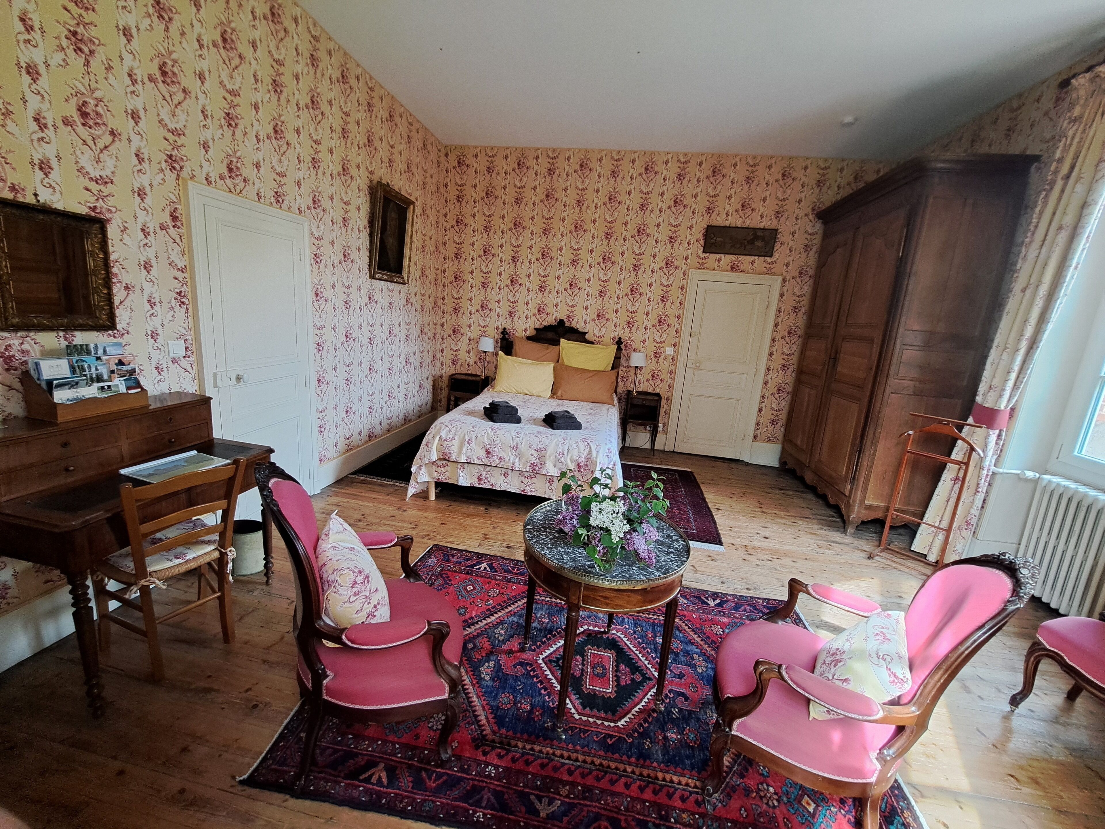 Una camera da letto con carta da parati floreale, un letto con cuscini gialli e neri, una scrivania in legno e due sedie rosa.