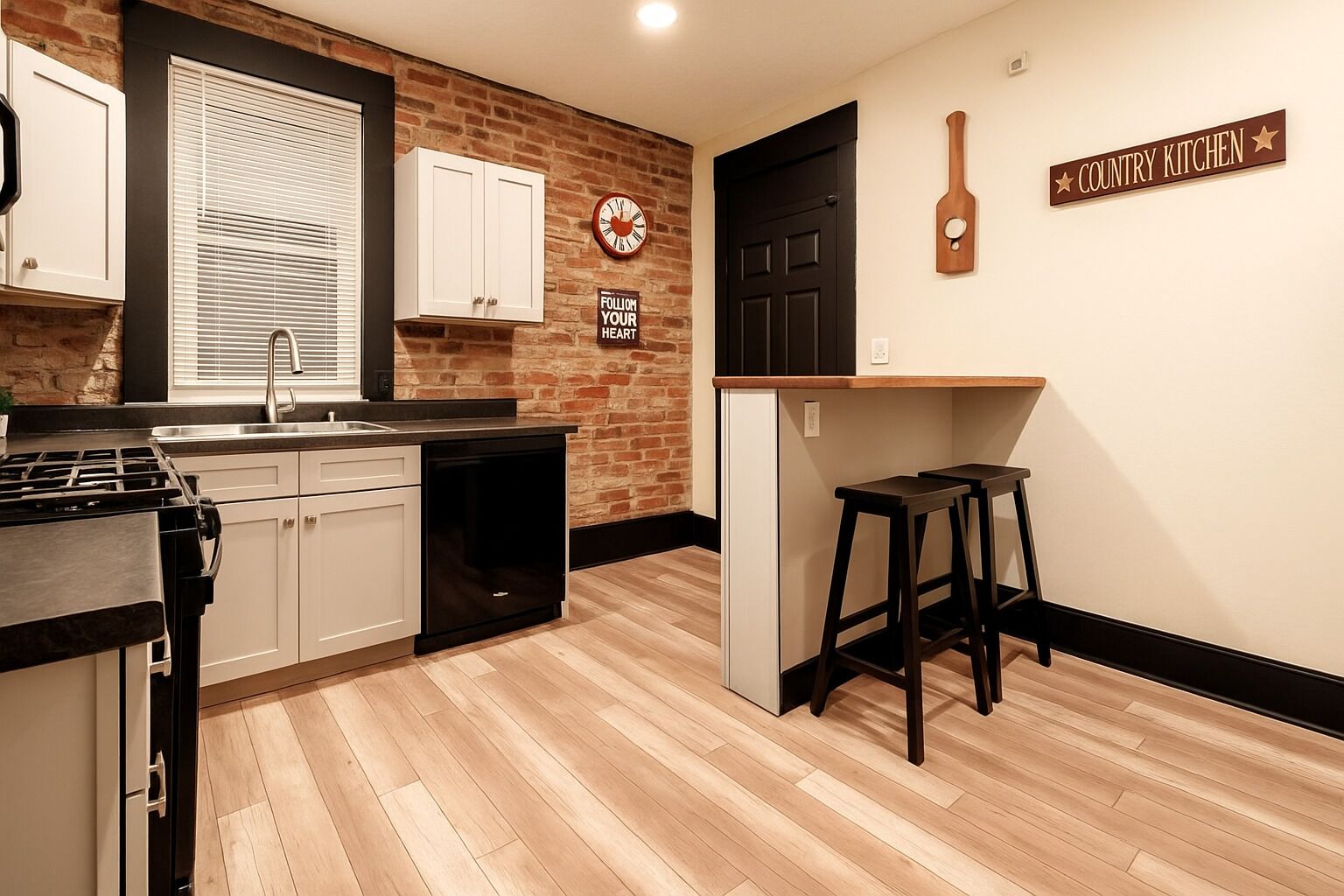 A kitchen with a brick wall, white cabinets, a black countertop, a stove, a dishwasher, a clock, and a sign that reads "Country Kitchen".