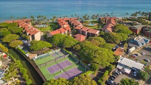 Una vista aerea di un resort costiero con diversi edifici, un campo da tennis e vista sull'oceano e sulle montagne.