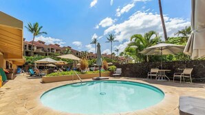Ein Swimmingpool mit Liegestühlen und Sonnenschirmen, umgeben von Palmen und Hotelgebäuden.