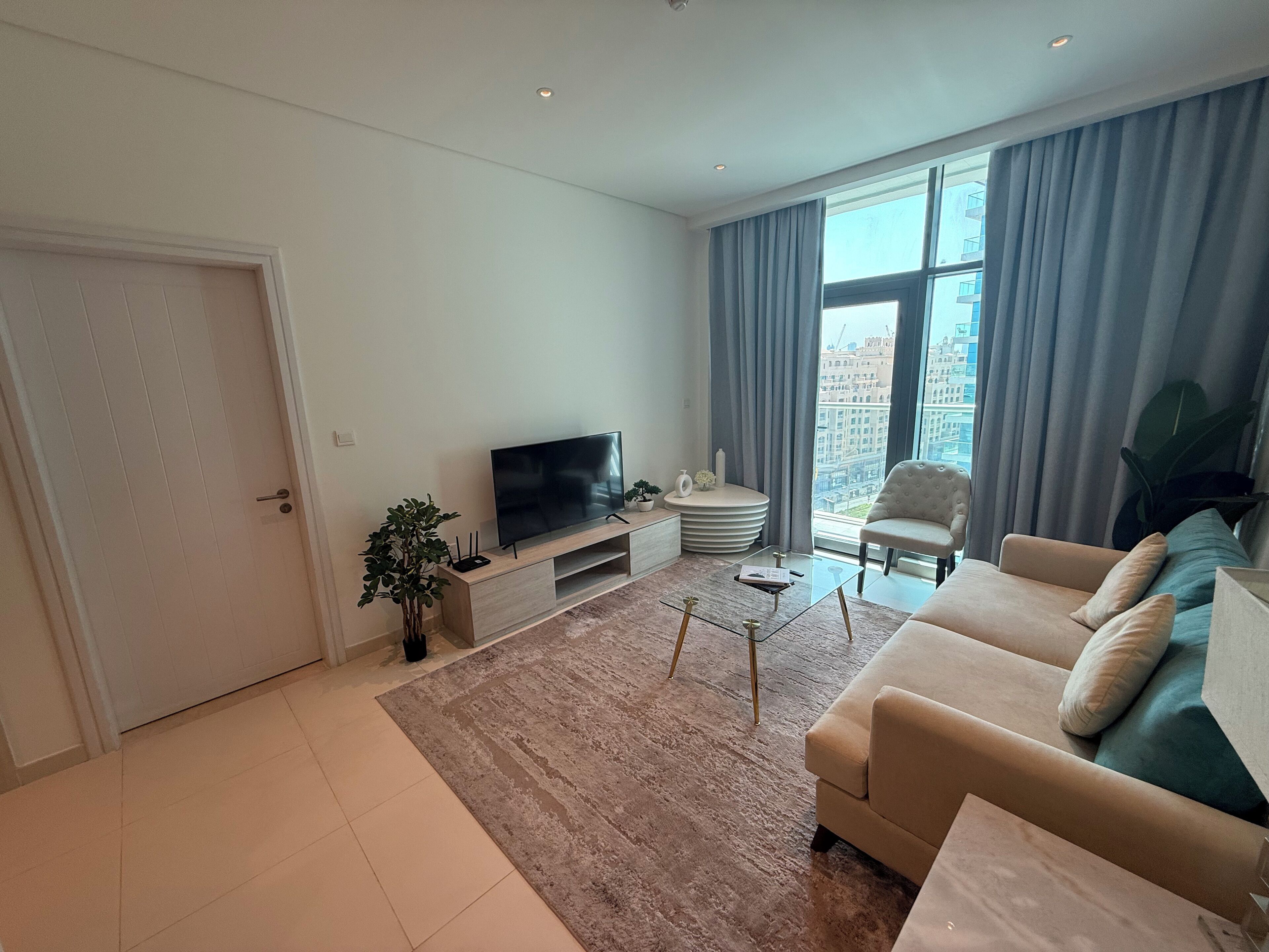 Een moderne woonkamer met een flatscreen televisie op een houten standaard, een beige bank, een glazen salontafel en een groot raam met gordijnen.