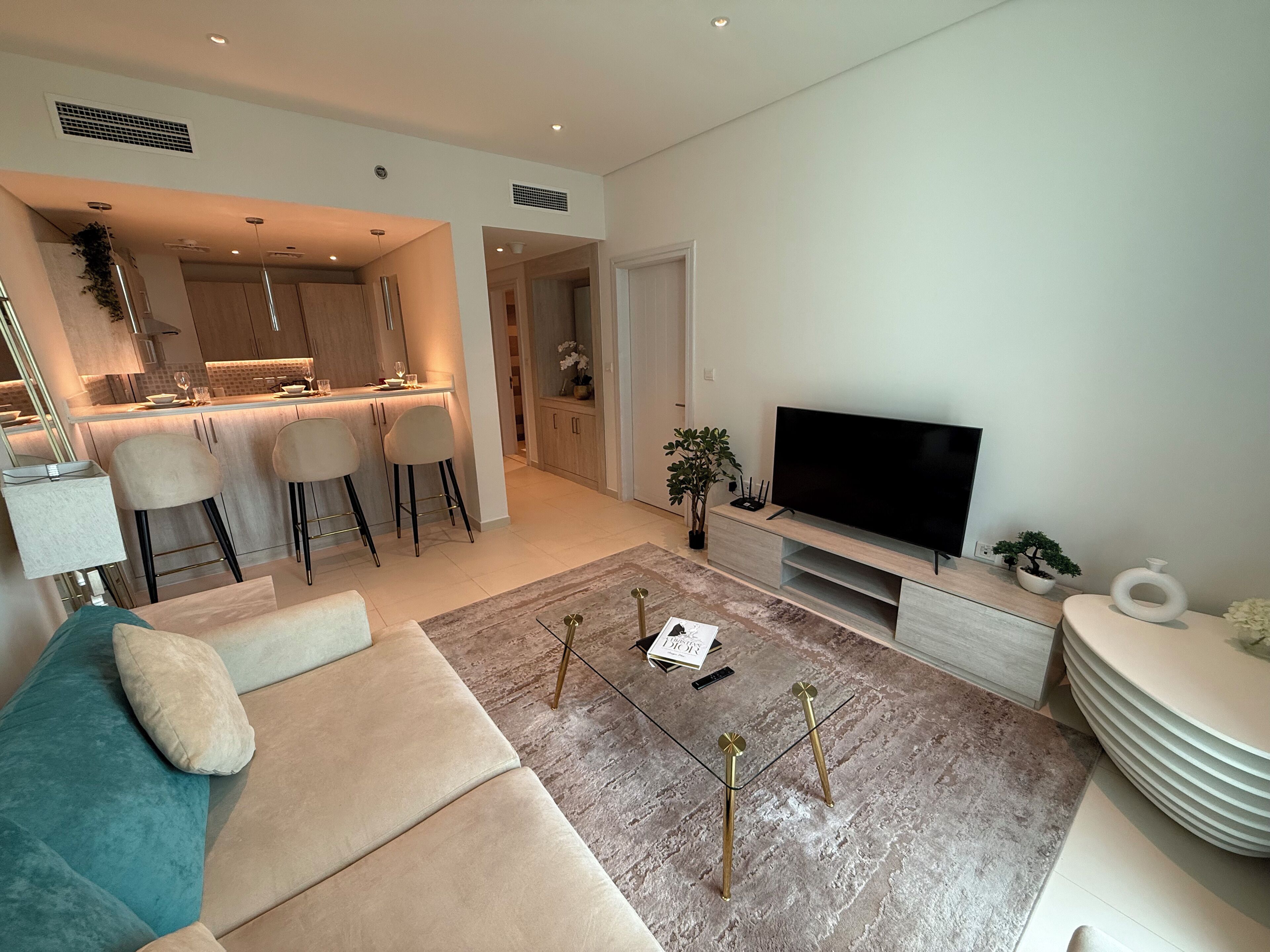 A modern living room with a sofa, a coffee table, and a TV. There is a kitchen area with a dining table and chairs.