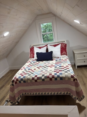 A bedroom with a sloped ceiling, a wooden floor, a bed with a quilt, a nightstand, and a window with a view of trees. - Pet-Friendly Table Rock Lake Cabin near Eureka Springs (Eagle Rock)