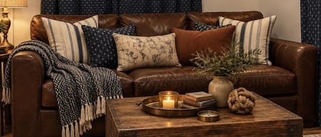 A cozy living room with a brown leather sofa, patterned cushions, a rustic wooden coffee table with a candle, and a blue curtain with a star pattern.