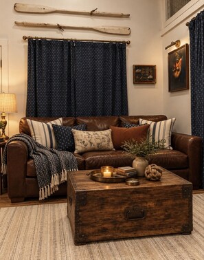 Una sala de estar acogedora con un sofá de cuero marrón, cojines estampados, una mesa de centro de madera rústica con una vela y una cortina azul con diseño de estrellas.