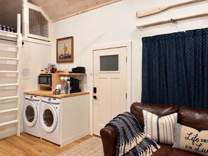 A compact living space with a washing machine, a microwave, a small kitchen area, a door, and a brown leather sofa with decorative pillows. - Pet-Friendly Table Rock Lake Cabin near Eureka Springs (Eagle Rock)