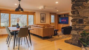A modern living room with a stone accent wall, a fireplace, a dining area with a table and chairs, and a large flat-screen TV displaying 'NETFLIX'.