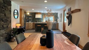 A dining area with a wooden table, chairs, and a vase with dried flowers. A kitchen is visible in the background.