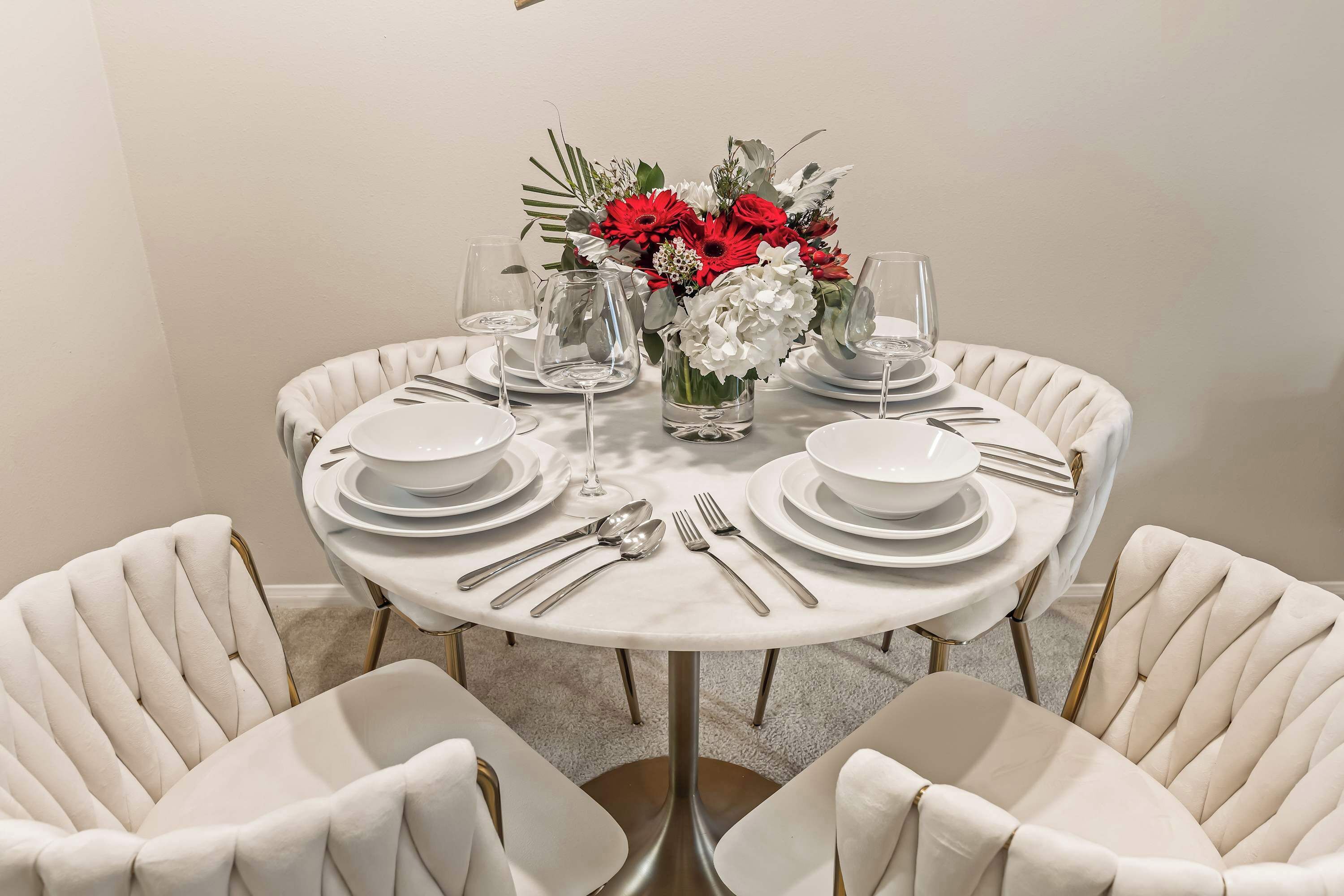 Une table ronde est dressée pour le dîner, avec de l’argenterie, de la vaisselle blanche et de la verrerie transparente. Un vase contenant des fleurs rouges et blanches est placé au centre.