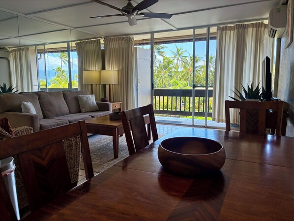 A living room with a sofa, dining table, chairs, a ceiling fan, and a view of palm trees through large windows.