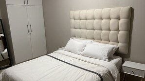 A bedroom with a large bed, white bedding, a tufted headboard, a wardrobe, and a bedside table.