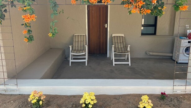 A covered outdoor seating area with two chairs, a wooden door, and a washing machine in the background.