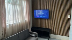 A modern living room with a flat-screen TV mounted on a wooden wall, a black sofa, and a window with sheer curtains.