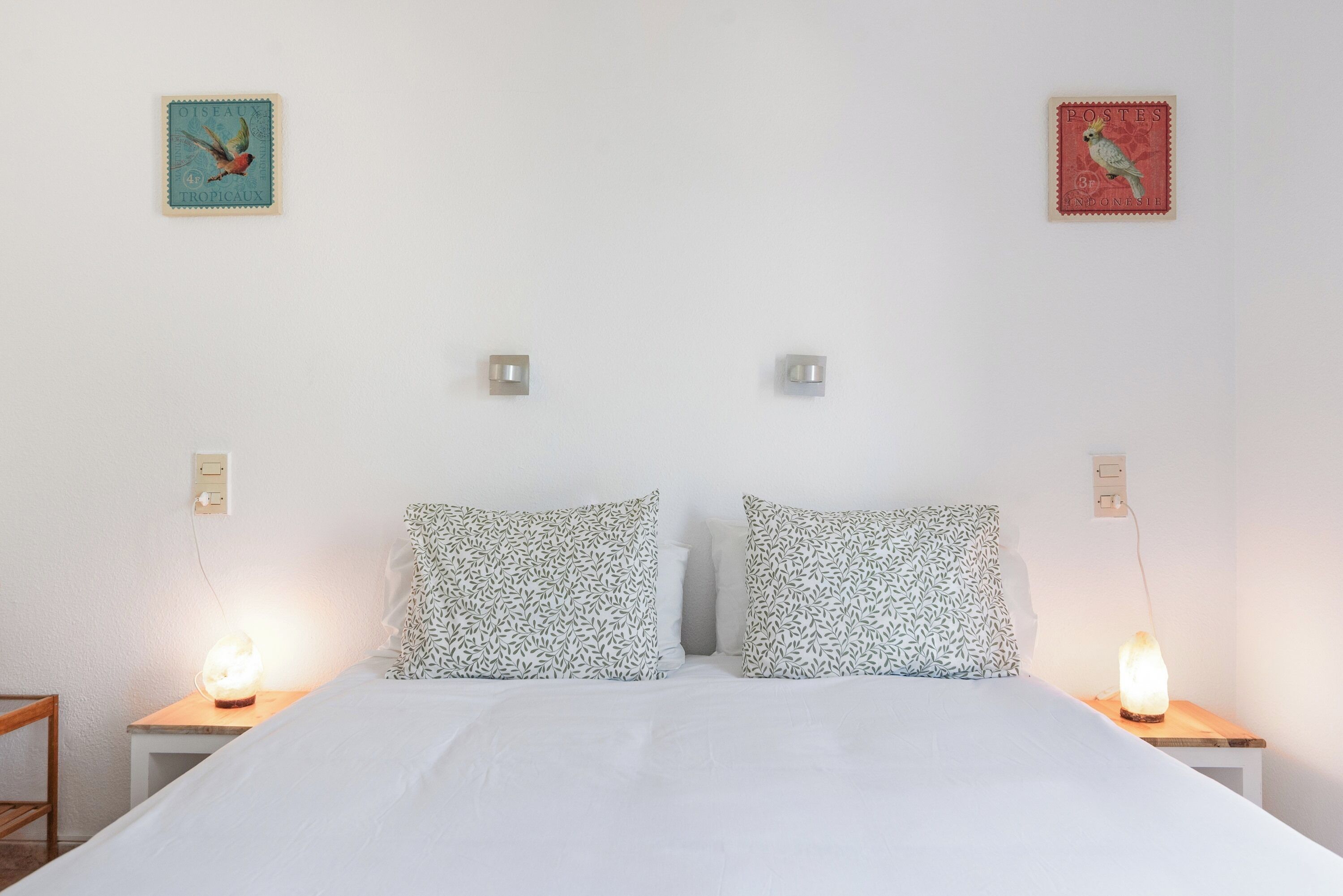 Un lit avec une literie blanche, deux coussins à motifs, deux lampes de chevet et deux interrupteurs d’éclairage fixés au mur.