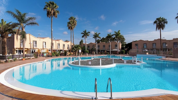 Une grande piscine aux eaux cristallines, entourée de palmiers et bordée d’une rangée de bâtiments beiges dotés de balcons.