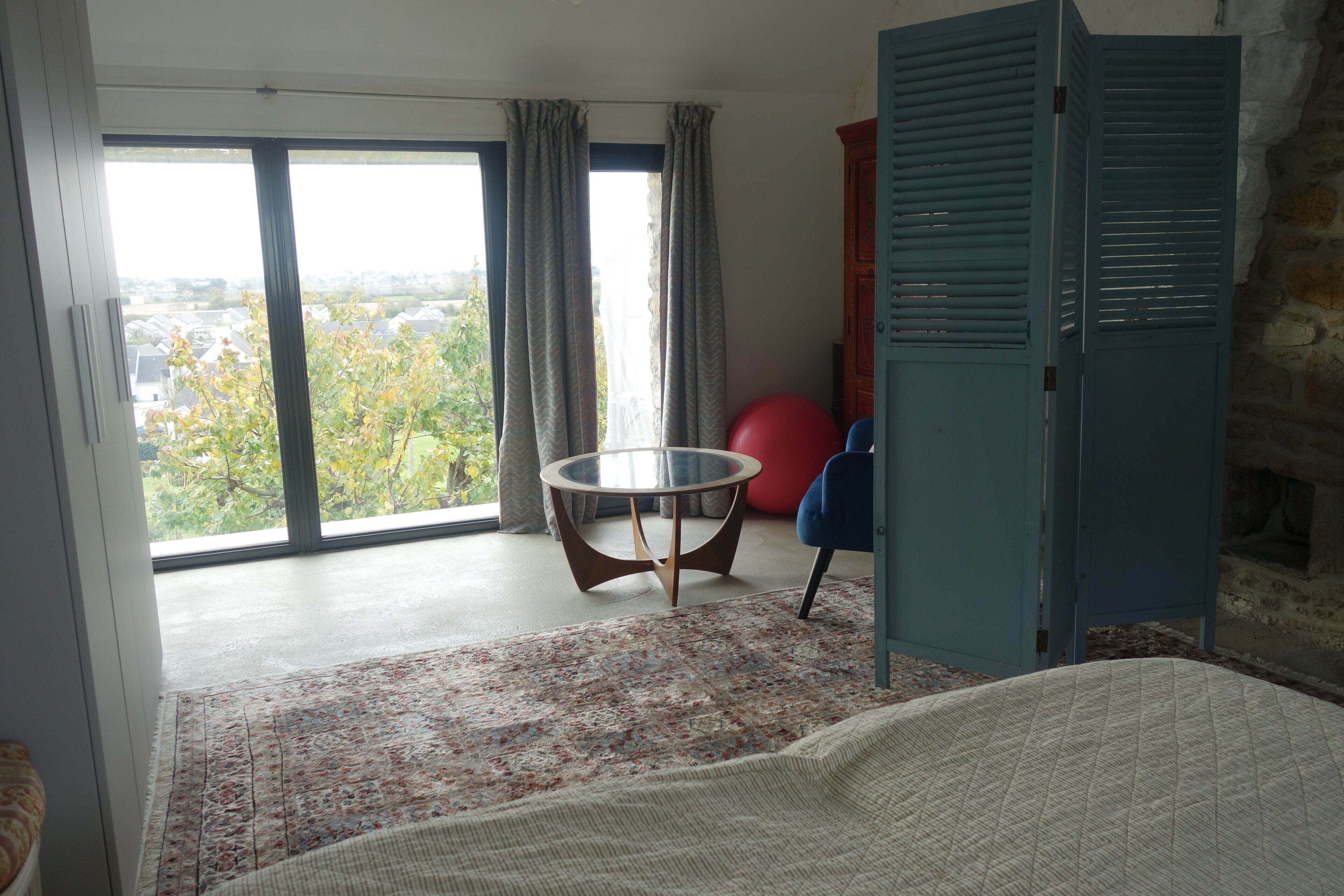 A room with a large window, a round table, a red exercise ball, a blue chair, and a wardrobe.