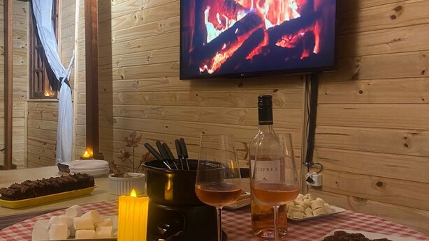 A cozy room with a wooden interior, a television displaying a fire, and a table set with snacks, wine, and a candle. - house with WiFi in Guarulhos you can share room and kitchen and hall with tv (Jardim Cristina Guarulhos)