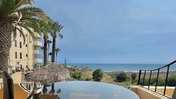 A round table with a glass top, surrounded by palm trees and a thatched roof structure, overlooking a beach and ocean. - Puerto Nuevo Villa Olas 2BR 2BA with pool (Centro Turístico El Pescador)