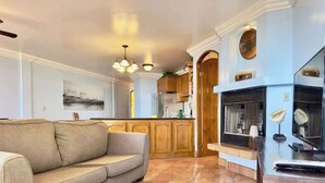 Sala de estar con sofá, una mesa de centro y chimenea. Hay una zona de cocina con armarios de madera y estufa.