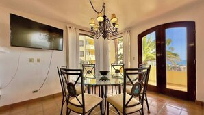 Un comedor con mesa de vidrio, cuatro sillas, una araña, un televisor de pantalla plana y vistas a palmeras a través de un ventanal.