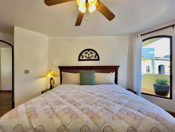 A bedroom with a large bed, a ceiling fan, a wall clock, and a window with a view of buildings.