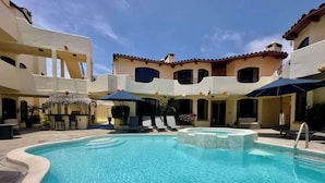 Una piscina con lettini e ombrelloni, circondata da edifici con balconi e un cielo sereno.