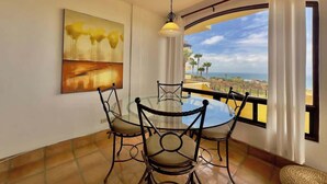 A dining area with a round table and chairs, a pendant light, a painting on the wall, and a view through large windows.