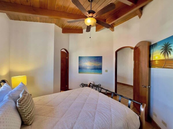 Un dormitorio con cama, ventilador de techo, pintura de atardecer y puertas de madera.