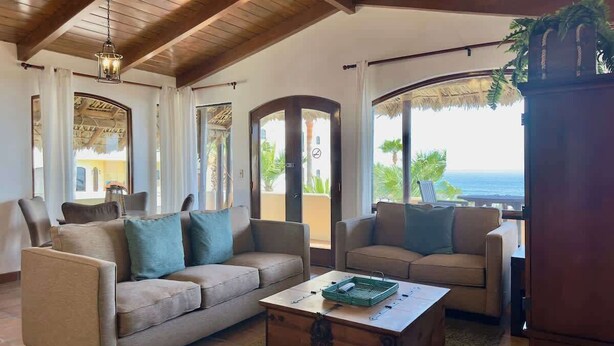 Amplia sala de estar con techo alto y vigas de madera, con dos sofás, una mesa de centro y grandes ventanales con vistas al océano.