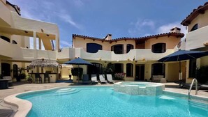 A swimming pool with lounge chairs and umbrellas, surrounded by buildings with balconies and a clear sky.