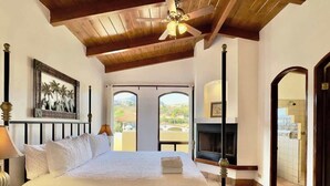 A bedroom with a bed, a ceiling fan, a fireplace, and a view of the outside.