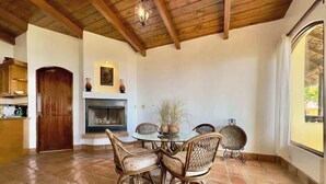 A dining area with a round table and wicker chairs, a fireplace, and a view through a window.