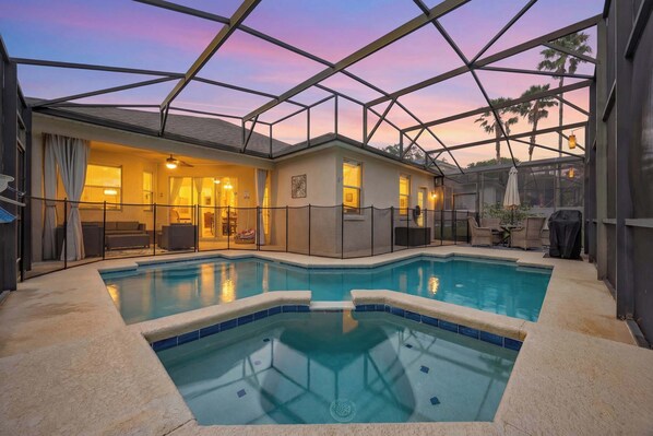 A swimming pool with a glass enclosure, a covered patio area, and a view of the sunset.