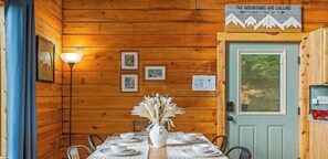 L’intérieur d’un chalet en bois, avec une table à manger dressée pour quatre personnes, un vase contenant des fleurs séchées, des tableaux encadrés accrochés au mur et une porte sur laquelle est apposée une pancarte.