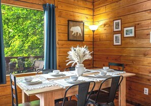 A wooden interior with a dining table set for four, a vase with dried plants, and framed pictures on the wall.