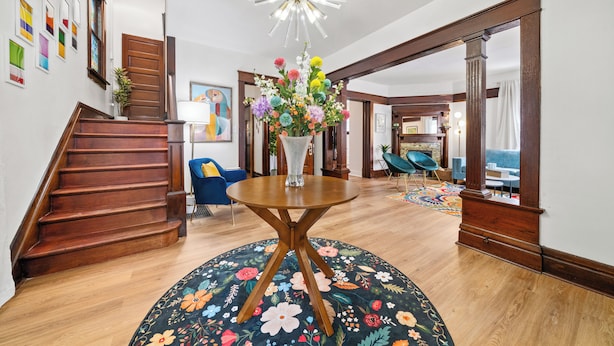 A staircase leading to a room with a wooden floor, a round table with a floral rug, and a chandelier.