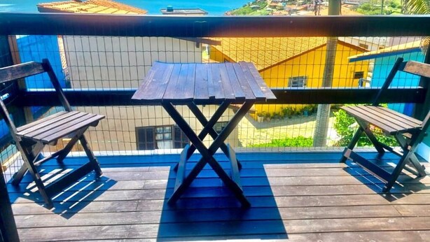 A wooden deck with a table and two chairs, overlooking a beach and a yellow building. - Chalés Santa Marta (Laguna)