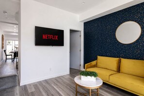 A modern living room with a flat-screen TV mounted on the wall, a yellow sofa, a round coffee table with a plant, and a dark speckled accent wall.
