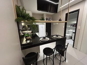 A compact kitchen with a black countertop, a TV above, and a small dining area with two chairs and a table set for two. - Bela Hospedagem - 607 One Way Tambau (Joao Pessoa)