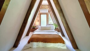 Een slaapkamer met een schuine kap, houten balken, een bed met wit beddengoed, een bruine plaid en een raam met uitzicht op groen.