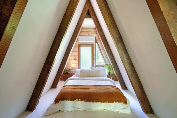 A bedroom with a sloped ceiling, wooden beams, a bed with white bedding, a brown throw blanket, and a window with a view of greenery outside. - Modern A-Frame - Hot Tub - Forest Escape (Rhododendron)